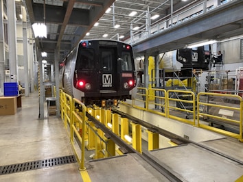 A metro train is positioned inside a maintenance facility. The train is elevated above the ground, supported by a track system, allowing for inspection or repairs. Various equipment and tools are visible around the area, with bright lighting illuminating the space. A worker is seen in the background, suggesting an active maintenance process.