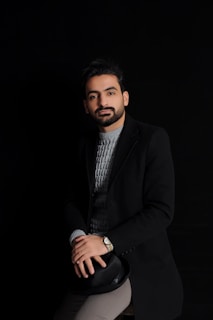 A well-groomed man with a beard is seated, wearing a dark coat over a gray knit sweater. He has a calm expression and is holding a black hat on his lap. The background is black, emphasizing a minimalist and sophisticated look.