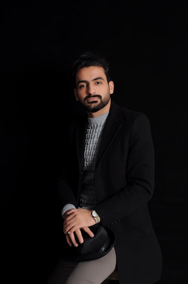 A well-groomed man with a beard is seated, wearing a dark coat over a gray knit sweater. He has a calm expression and is holding a black hat on his lap. The background is black, emphasizing a minimalist and sophisticated look.