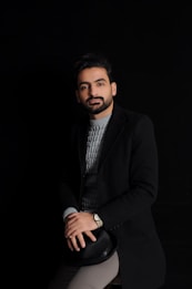 A well-groomed man with a beard is seated, wearing a dark coat over a gray knit sweater. He has a calm expression and is holding a black hat on his lap. The background is black, emphasizing a minimalist and sophisticated look.