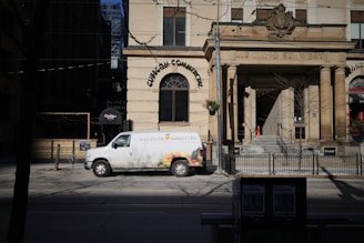 A white van with 'Augusta Fruit Market Ltd' written on the side is parked on the street in front of a historic building with large columns. The building has an inscription '212 King St. West' above the entrance. There are some trees and a street sign nearby, with the shadow of a leafless tree cast on the street.