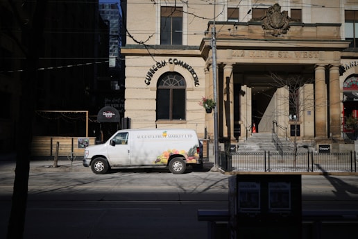 A white van with 'Augusta Fruit Market Ltd' written on the side is parked on the street in front of a historic building with large columns. The building has an inscription '212 King St. West' above the entrance. There are some trees and a street sign nearby, with the shadow of a leafless tree cast on the street.