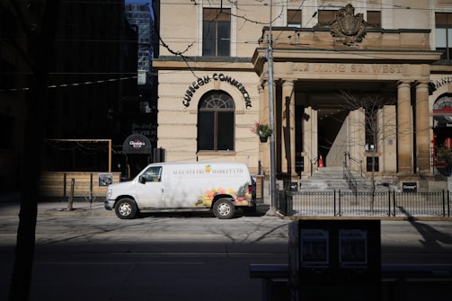 A white van with 'Augusta Fruit Market Ltd' written on the side is parked on the street in front of a historic building with large columns. The building has an inscription '212 King St. West' above the entrance. There are some trees and a street sign nearby, with the shadow of a leafless tree cast on the street.