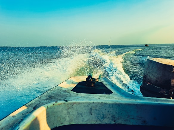 A motorboat speeds through the open water, creating a trail of frothy white waves. The clear blue sky stretches above, and the horizon is visible in the distance. The water reflects various shades of blue and green, accentuating the movement of the boat.