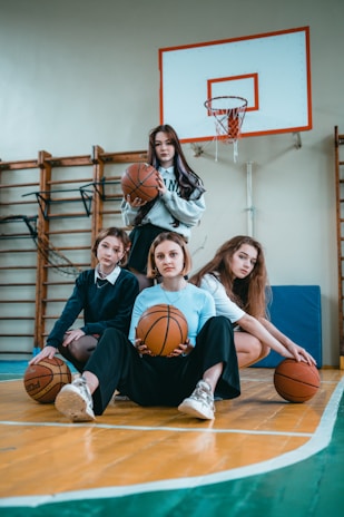 Four young women are posing on a basketball court, each holding a basketball. The background includes a basketball hoop and gym equipment, indicating an indoor sports facility. Their expressions and postures suggest camaraderie and confidence.