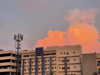 A multi-story building stands prominently against a backdrop of vibrant orange clouds during sunset. An adjacent telecommunications tower is visible on the left. The sky has a gentle gradient from the warm cloud colors to a cooler gray-blue.