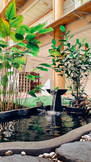 A small indoor water fountain features gently flowing water surrounded by lush green plants. The area is enclosed in a modern architectural setting with high ceilings and visible structural elements. Smooth pebbles are scattered around the fountain's edge, and there is a subtle play of light on the water surface creating reflections.