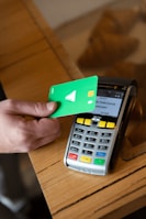 A hand holds a green debit card near a payment terminal ready for a contactless transaction. The terminal displays various function buttons and an LCD screen showing transaction options.