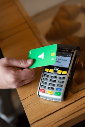 A hand holds a green debit card near a payment terminal ready for a contactless transaction. The terminal displays various function buttons and an LCD screen showing transaction options.