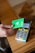 A hand holds a green debit card near a payment terminal ready for a contactless transaction. The terminal displays various function buttons and an LCD screen showing transaction options.