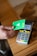 A hand holds a green debit card near a payment terminal ready for a contactless transaction. The terminal displays various function buttons and an LCD screen showing transaction options.