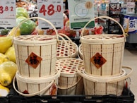 A stack of woven bamboo baskets with handles, displayed in a market. Each basket has a red diamond-shaped label featuring Chinese characters. Surrounding the baskets are signs indicating prices in various currencies and informational signage mostly in Chinese. To the left, there are green and yellow oversized pomelos, adding to the market setting.