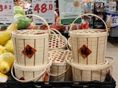 A stack of woven bamboo baskets with handles, displayed in a market. Each basket has a red diamond-shaped label featuring Chinese characters. Surrounding the baskets are signs indicating prices in various currencies and informational signage mostly in Chinese. To the left, there are green and yellow oversized pomelos, adding to the market setting.