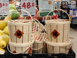 A stack of woven bamboo baskets with handles, displayed in a market. Each basket has a red diamond-shaped label featuring Chinese characters. Surrounding the baskets are signs indicating prices in various currencies and informational signage mostly in Chinese. To the left, there are green and yellow oversized pomelos, adding to the market setting.