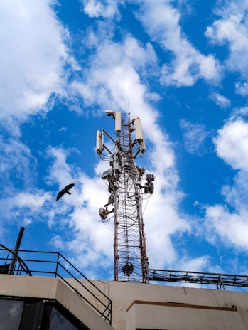 A tall communication tower laden with various antennas and equipment stands against a vivid blue sky scattered with white clouds. Below the tower, part of a building's roof with railings is visible. A bird is in flight near the tower, providing a sense of scale and motion to the scene.