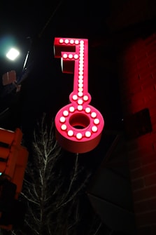 A large, illuminated red key sign is mounted against a dark background. The key is adorned with bright white lights outlining its shape, creating a striking contrast against the night sky. A dimly lit streetlight and a tree branch are visible in the background, adding to the urban setting.