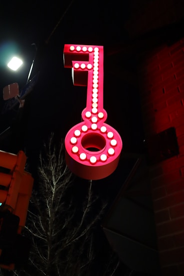 A large, illuminated red key sign is mounted against a dark background. The key is adorned with bright white lights outlining its shape, creating a striking contrast against the night sky. A dimly lit streetlight and a tree branch are visible in the background, adding to the urban setting.