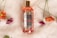 A bottle of bath and shower gel labeled Skyn & Shower is placed on a soft, fluffy white surface. The label includes floral decorations in red, pink, and green. Surrounding the bottle are several vibrant flowers in shades of orange and purple, with green stems and leaves.