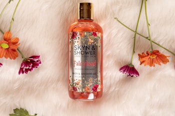 A bottle of bath and shower gel labeled Skyn & Shower is placed on a soft, fluffy white surface. The label includes floral decorations in red, pink, and green. Surrounding the bottle are several vibrant flowers in shades of orange and purple, with green stems and leaves.