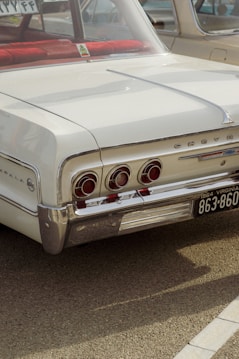 A vintage Chevrolet Impala is parked with its focus on the rear section. The car is white with chrome detailing and features traditional round taillights. The license plate is from Virginia, dated 1964. A red blanket or item is visible through the rear window.