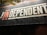 A sign with bold text reads 'THE INDEPENDENT' with a stylized emphasis on 'IN'. The background is primarily dark, creating a contrast with the light-colored text. There are some overhanging leaves visible at the top.