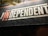 A sign with bold text reads 'THE INDEPENDENT' with a stylized emphasis on 'IN'. The background is primarily dark, creating a contrast with the light-colored text. There are some overhanging leaves visible at the top.