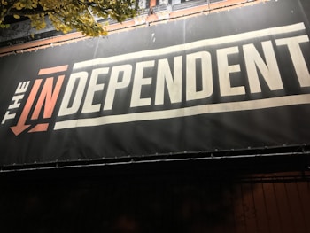 A sign with bold text reads 'THE INDEPENDENT' with a stylized emphasis on 'IN'. The background is primarily dark, creating a contrast with the light-colored text. There are some overhanging leaves visible at the top.