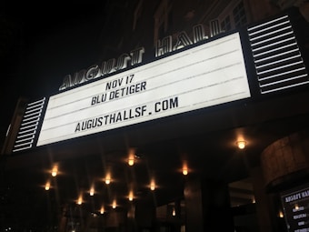 An illuminated theater marquee displays event information for an upcoming performance by Blu DeTiger on November 17 at August Hall. Surrounding the marquee are a series of lit bulbs, creating a warm, inviting atmosphere.