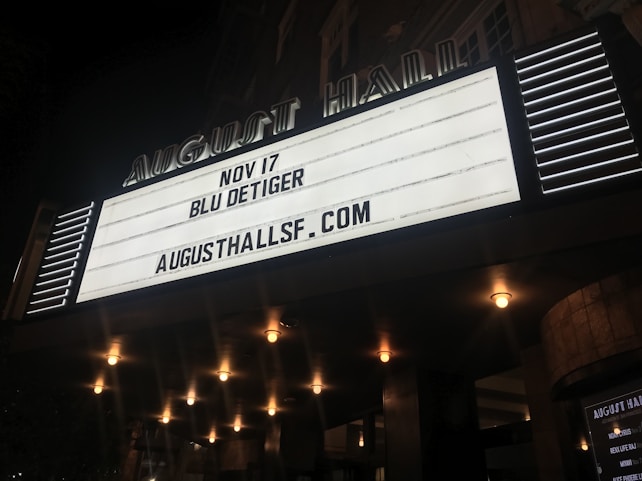 An illuminated theater marquee displays event information for an upcoming performance by Blu DeTiger on November 17 at August Hall. Surrounding the marquee are a series of lit bulbs, creating a warm, inviting atmosphere.