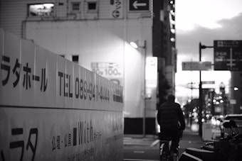 An urban street scene captured in black and white, with a cyclist wearing a dark jacket riding along a city block. The side of a building is visible with Japanese text and a phone number displayed prominently. The background features several signs and a slightly blurred cityscape as evening falls, conveyed by the lowered light and shadows.