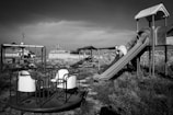 An abandoned playground with a slide, a merry-go-round, and other playground equipment. The area is overgrown with grass and weeds, and the structures appear weathered and unused. There are buildings and structures in the background, with a generally desolate atmosphere.