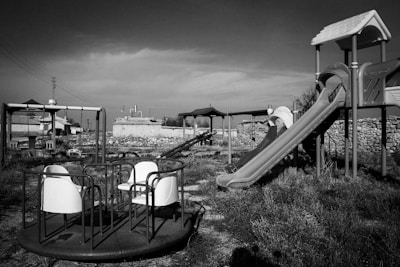 An abandoned playground with a slide, a merry-go-round, and other playground equipment. The area is overgrown with grass and weeds, and the structures appear weathered and unused. There are buildings and structures in the background, with a generally desolate atmosphere.