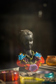 A dark mannequin bust displays an intricately designed necklace featuring vibrant flowers in shades of blue, red, purple, and orange. Surrounding the bust are various colorful glass objects, including bracelets and small decorative bowls.