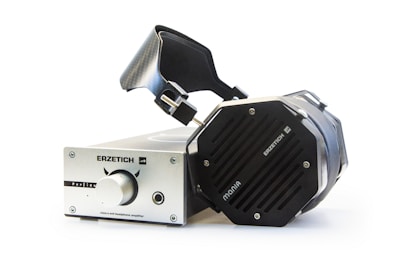 A pair of large, premium over-ear headphones connected to a high-end headphone amplifier. The headphones feature a robust, angular design with visible metal components, while the amplifier has a metallic finish with a central knob and labeled controls.