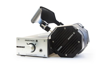 A pair of large, premium over-ear headphones connected to a high-end headphone amplifier. The headphones feature a robust, angular design with visible metal components, while the amplifier has a metallic finish with a central knob and labeled controls.