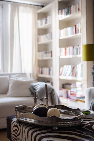 A cozy and modern living room features a tray with two candles and decorative balls placed on a zebra-patterned ottoman. Surrounding the space are large bookshelves filled with various colored books, contributing to the room’s vibrant and intellectual atmosphere. Soft natural light filters in through sheer curtains, illuminating a comfortable white sofa.