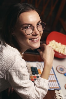 A person wearing glasses and a white textured shirt is smiling while sitting at a table. In front of them, there is a colorful board game with tiles and a red bowl filled with popcorn.