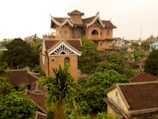 A large, intricately designed house with Asian architectural influences is surrounded by lush greenery. The building features a distinctive roof style, arched entryways, and round windows. The scene is set in a serene, rural environment with additional smaller buildings visible among the trees.