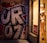 A large piece of bold graffiti with stylized letters is painted on an urban wall. Next to it, a shop window displays a mannequin wearing lingerie. A red 'SALE' sign is visible, and the scene is illuminated by warm lighting.