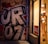 A large piece of bold graffiti with stylized letters is painted on an urban wall. Next to it, a shop window displays a mannequin wearing lingerie. A red 'SALE' sign is visible, and the scene is illuminated by warm lighting.