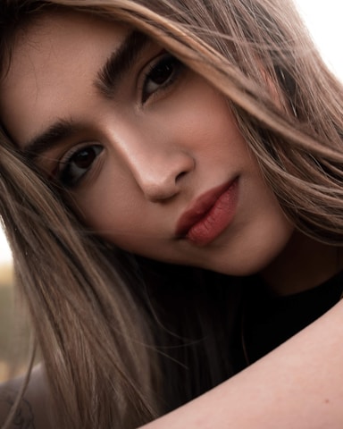 A close-up portrait of a young person with long hair, showcasing natural makeup and strong eyebrows against a soft, blurred background.