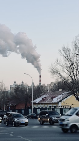 A cityscape with vehicles on a busy road, buildings in the background, and a prominent smokestack emitting thick clouds of smoke into the sky. Leafless trees are visible, and the sky is clear with a pale blue hue.