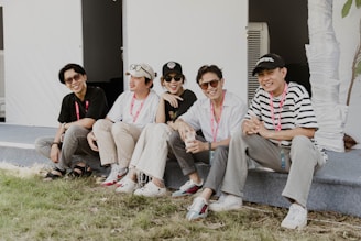 Five individuals are sitting on an outdoor ledge, wearing casual clothing and sunglasses, each with lanyards around their necks. They appear relaxed and are smiling, suggesting a cheerful group atmosphere. The background features a white structure with an air conditioning unit visible behind them.