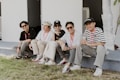 Five individuals are sitting on an outdoor ledge, wearing casual clothing and sunglasses, each with lanyards around their necks. They appear relaxed and are smiling, suggesting a cheerful group atmosphere. The background features a white structure with an air conditioning unit visible behind them.