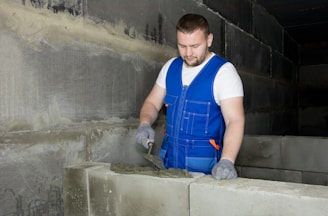 A man wearing a blue work vest and gloves is applying mortar on a row of concrete blocks in what appears to be a construction site. The walls around are made of similar blocks, and the environment looks industrial.
