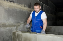 A man wearing a blue work vest and gloves is applying mortar on a row of concrete blocks in what appears to be a construction site. The walls around are made of similar blocks, and the environment looks industrial.