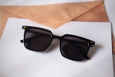 A pair of black sunglasses with square frames rests on top of some overlapping sheets of paper and a brown envelope. The sunglasses have a glossy finish with small gold accents on the corners.