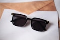 A pair of black sunglasses with square frames rests on top of some overlapping sheets of paper and a brown envelope. The sunglasses have a glossy finish with small gold accents on the corners.