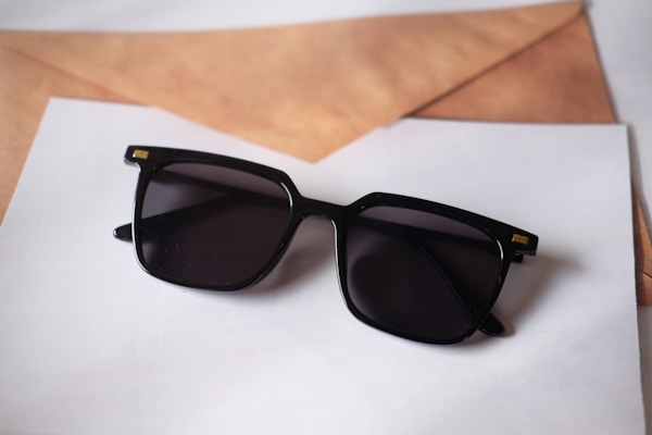 A pair of black sunglasses with square frames rests on top of some overlapping sheets of paper and a brown envelope. The sunglasses have a glossy finish with small gold accents on the corners.