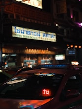 A street scene at night with a taxi in the foreground displaying a light-up 'TAXI-METER' sign. The setting appears to be an urban Chinatown with a large illuminated sign in Thai and English reading 'CHINA TOWN SCALA'. The surrounding area is dimly lit with a mix of colorful lights.